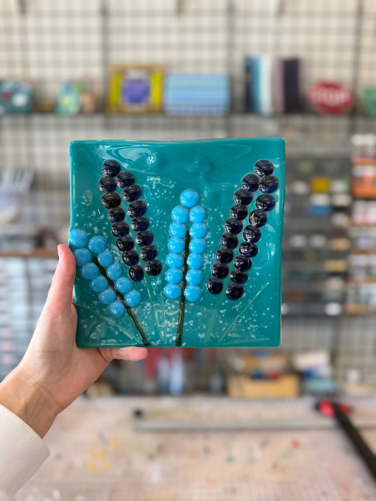 A hand holding a teal fused glass square decorated with blue glass dots at Paint Fusion Park City